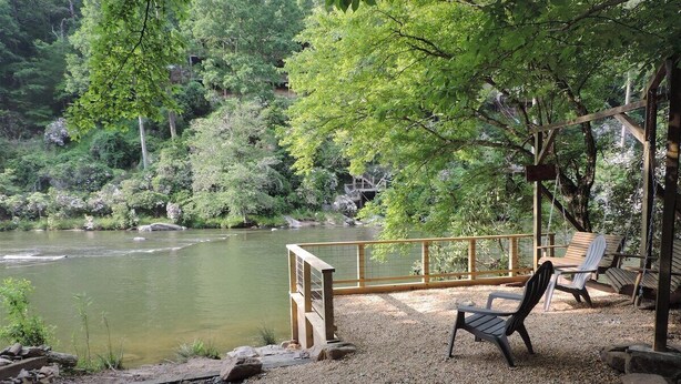 A riverside seating area with a wooden deck, chairs, and a view of a river surrounded by trees. - River Daze - A Beautiful River Front Brick House O (Ellijay)