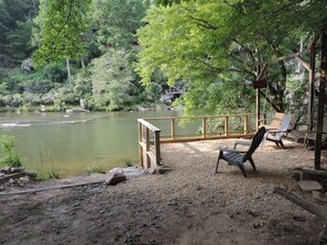A riverside seating area with a wooden deck, chairs, and a view of a river surrounded by trees.