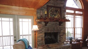 A cozy living room with a stone fireplace, a cushioned chair, a lamp, and a large window. - River Daze - A Beautiful River Front Brick House O (Ellijay)