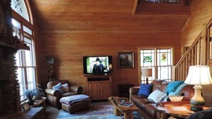 A cozy log cabin interior with a fireplace, leather sofas, a television, and a wooden staircase. - River Daze - A Beautiful River Front Brick House O (Ellijay)