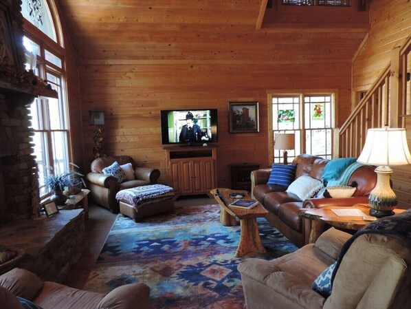 A cozy log cabin interior with a fireplace, leather sofas, a television, and a wooden staircase.