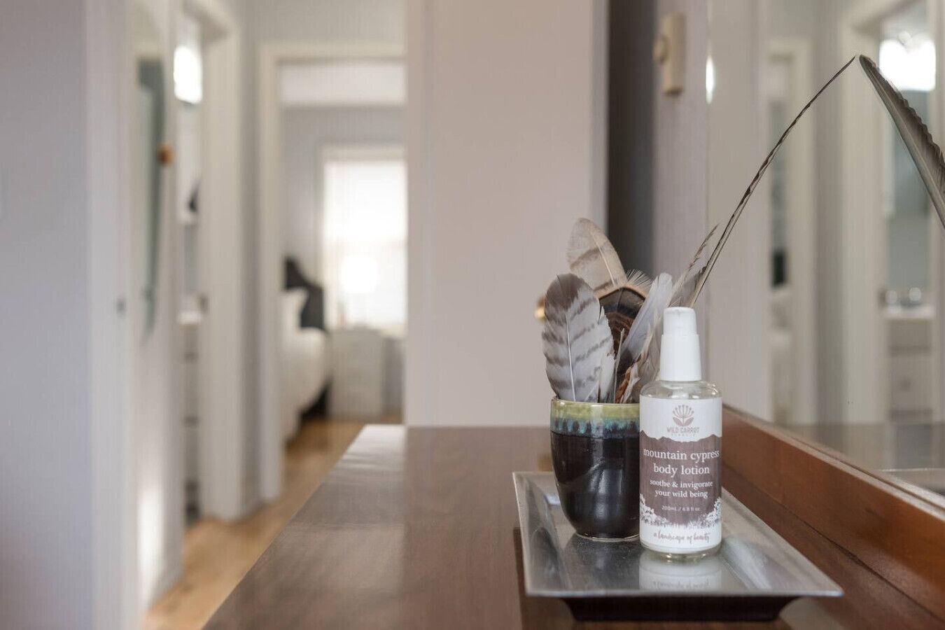 A bathroom with a wooden vanity, a mirror, and a bottle of Wild Carrot mountain cypress body lotion.