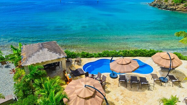 Un espace piscine situé au bord de la plage, agrémenté de chaises longues et de parasols, entouré d’une végétation luxuriante et offrant une vue dégagée sur l’océan.