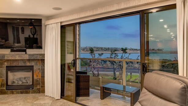 A modern living room with a fireplace, a comfortable sofa, and a large sliding glass door leading to a balcony with a view of the sea. - OCEANFRONT LUXURY—Summer booking fast so Hurry and book!<br> (San Diego)