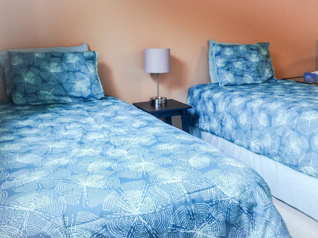 Ein Zimmer mit zwei Betten, jedes mit einer blauen, gemusterten Tagesdecke, einem kleinen Nachttisch mit einer Lampe und einer schlichten Wand.