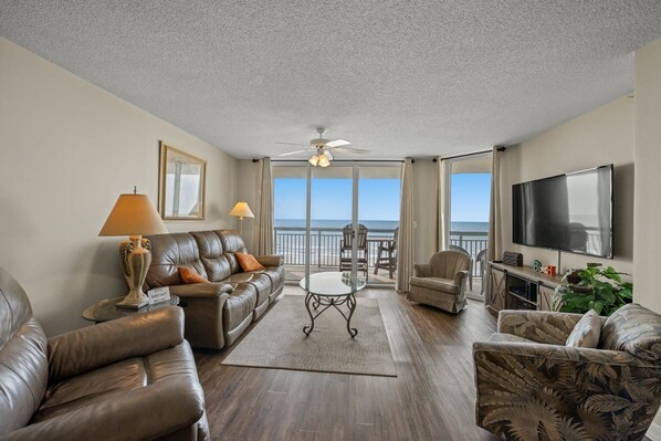 A living room with leather sofas, a glass coffee table, a TV, and a balcony with a view of the ocean.