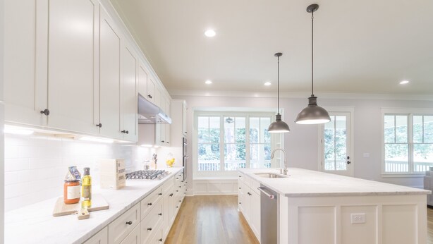 Cocina con armarios blancos, una isla central, iluminación colgante y vistas al exterior a través de amplios ventanales.
