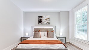 A modern bedroom with a wooden bed, white walls, and a large window with blinds. - Beautiful New Construction Home in Buckhead! Convenience & Luxury! (Atlanta)