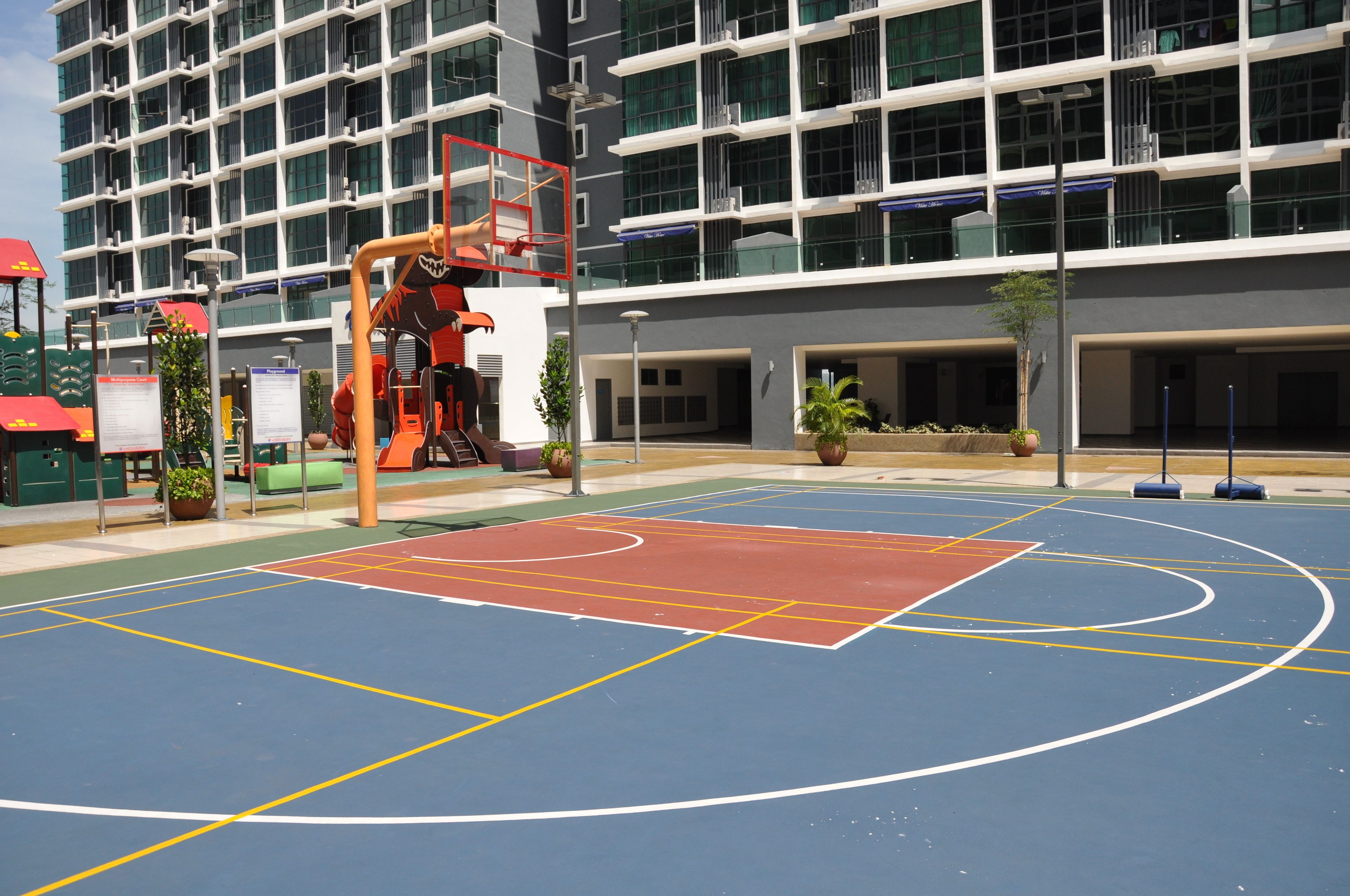 Un terrain de basket en plein air avec une surface de jeu rouge et bleue, entouré d’un bâtiment moderne doté de nombreux balcons et fenêtres.
