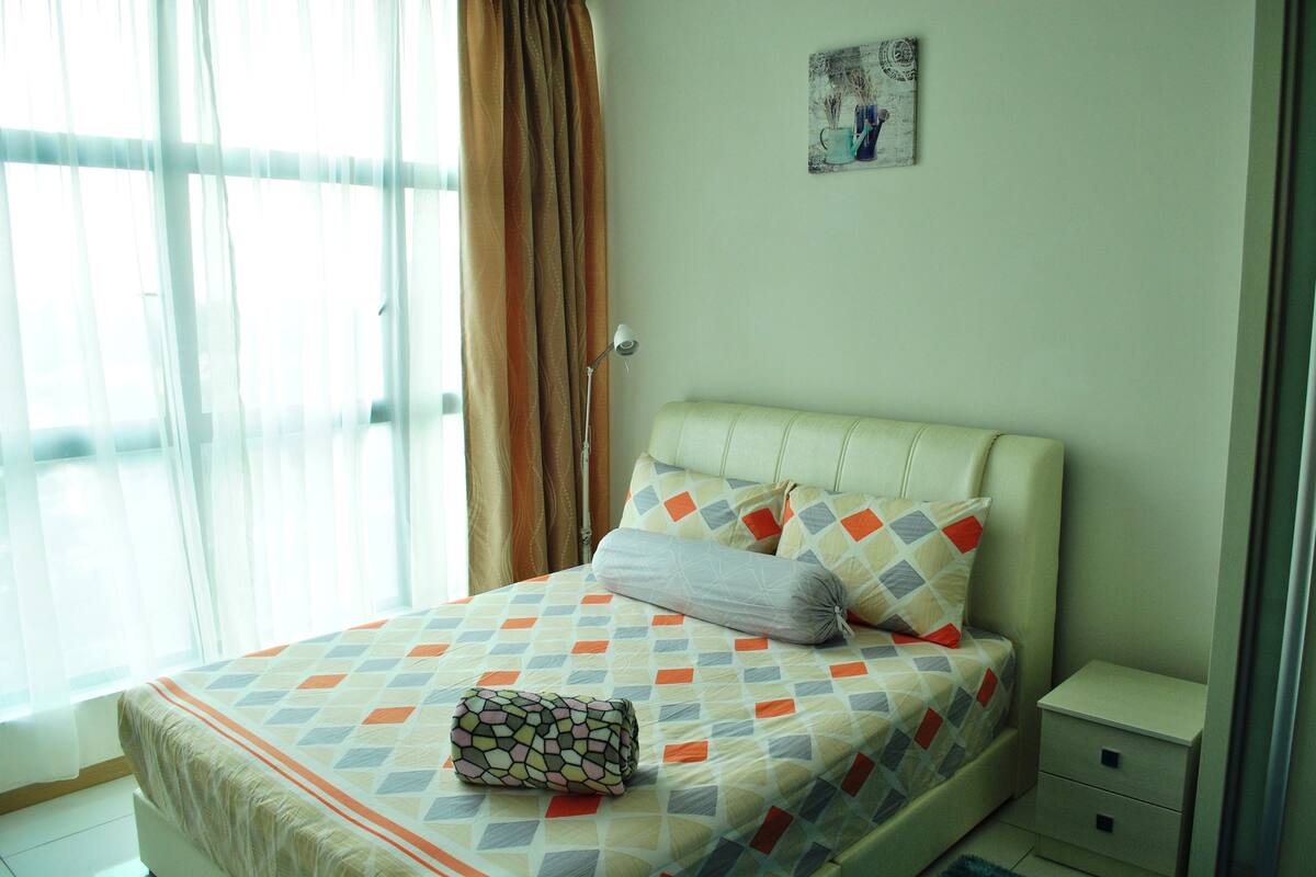 A neatly made bed with a patterned bedspread and pillows, a bedside table, and a window with curtains.