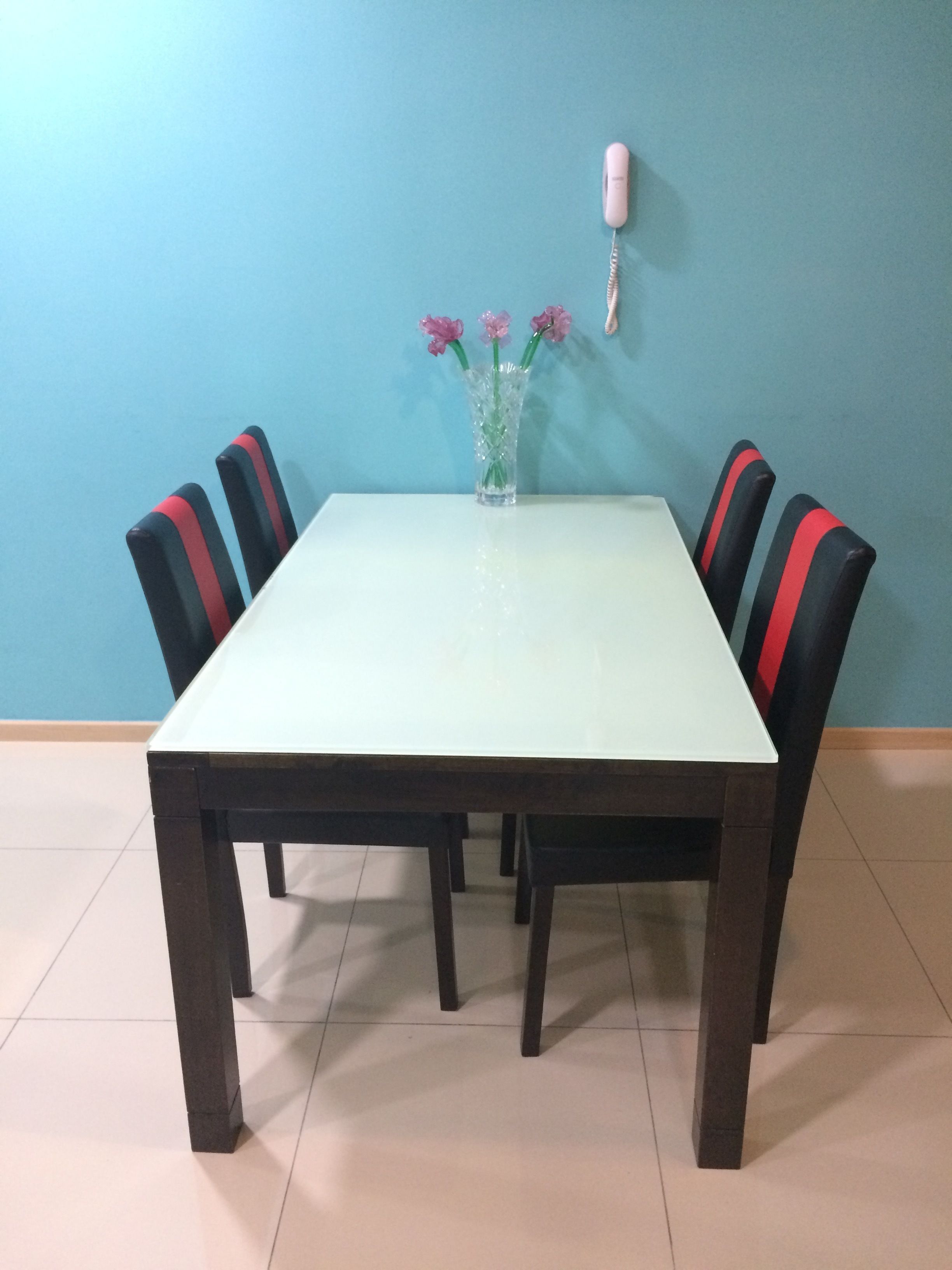 Une table de salle à manger rectangulaire, dotée d’un plateau blanc et de pieds foncés, est entourée de six chaises à rayures rouges et noires. Un vase contenant des fleurs roses est posé sur la table.