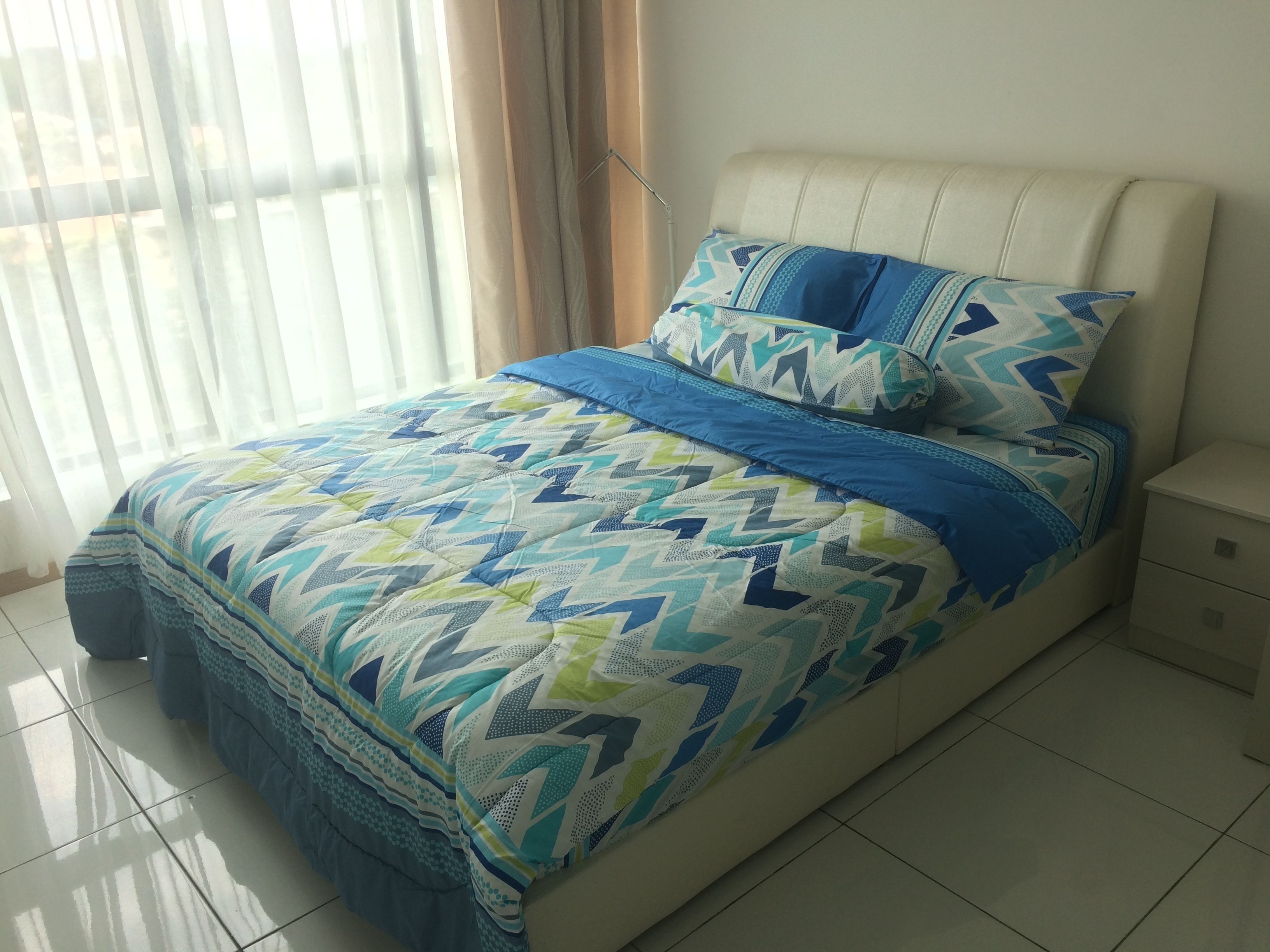 Una cama con colcha y almohadas de estampado geométrico, una mesita de noche y una ventana con cortinas translúcidas.