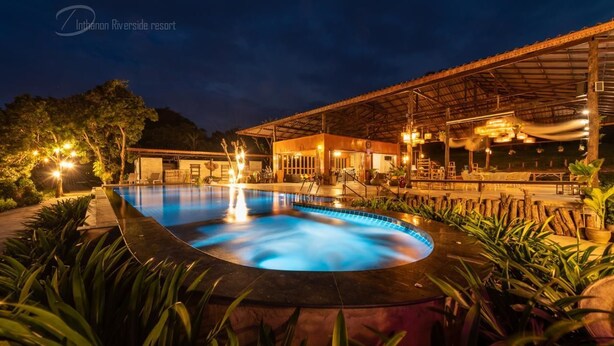 A swimming pool with a curved edge, surrounded by lush greenery and a covered outdoor area with seating and lighting. - Doi Inthanon Riverside Resort (Wiang Nong Long)