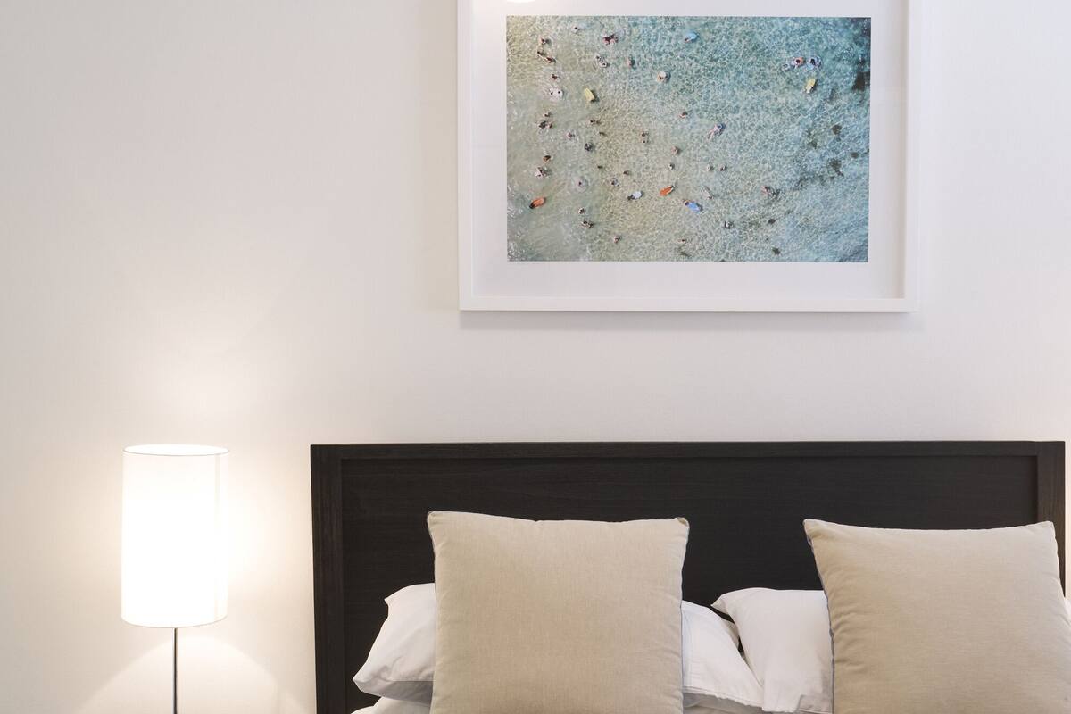 A neatly made bed with a dark headboard, a framed artwork above it, and a bedside lamp on a small table.