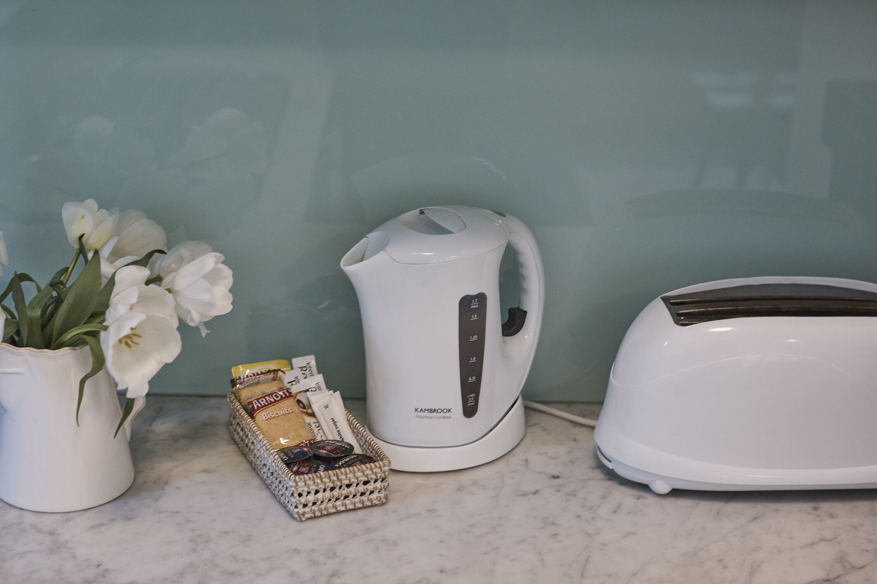 A kitchen counter with a white electric kettle, a toaster, a vase with white flowers, and a basket of snacks.