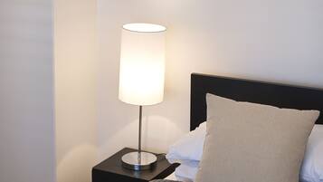 A neatly made bed with a grey quilt, a bedside lamp, and a pillow on a black nightstand.