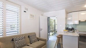 Una sala de estar moderna con un sofá color beige, cojines estampados, una cocina con armarios blancos y una pequeña isla con un cuenco de frutas.