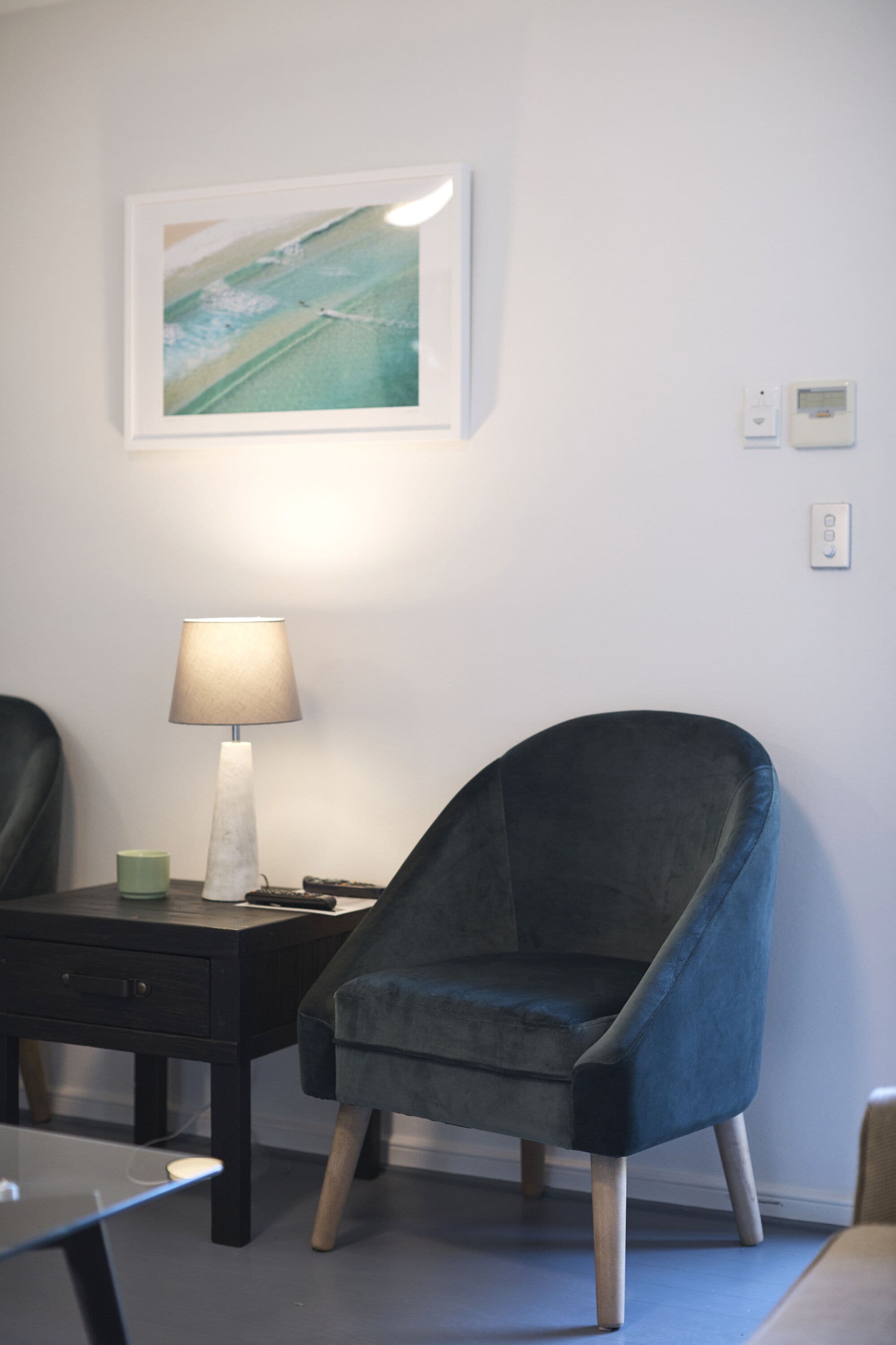A room with a dark blue chair, a small table with a lamp, and a framed picture on the wall.