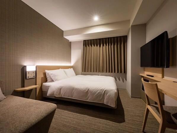 Ein Hotelzimmer mit einem großen Bett, einem Schreibtisch, einem Sessel, einem Fernseher und einer wandmontierten Steuerung.