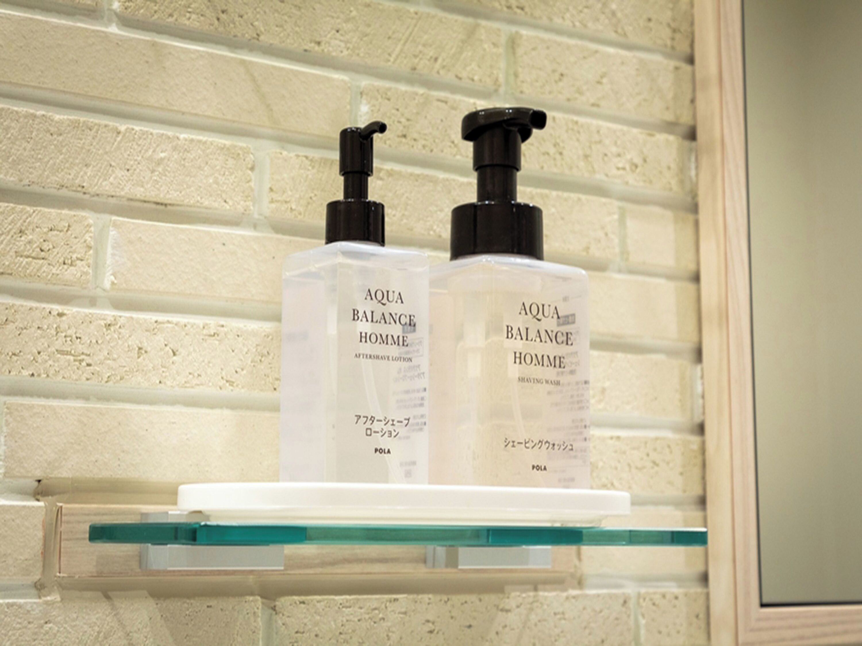 Two bottles of Aqua Balance Homme on a glass shelf against a white brick wall.