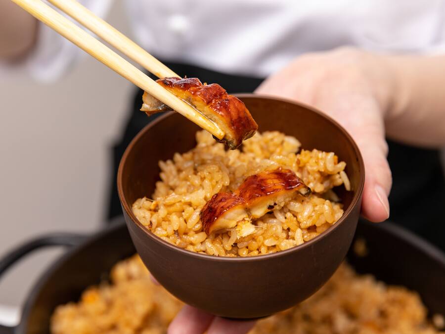 A bowl of rice with a piece of meat being held by chopsticks.