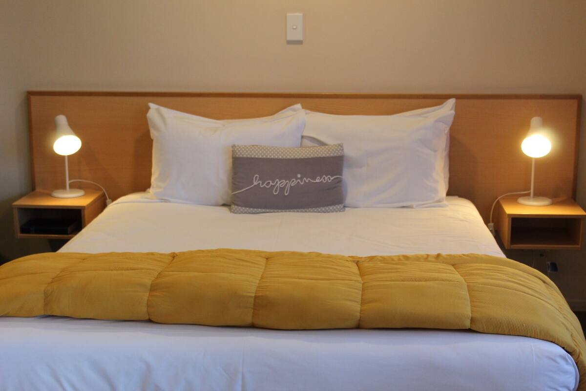 A neatly made bed with a yellow blanket, two bedside lamps, and a pillow with the word "joy" on it.