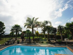 A swimming pool with surrounding palm trees and outdoor furniture. - ASURE Kaimai View Motel (Katikati)