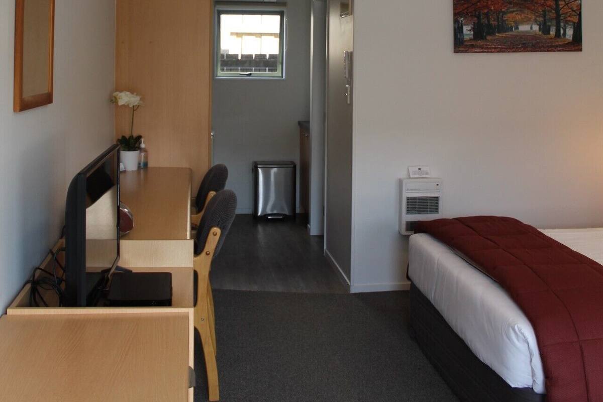 A compact hotel room with a bed, a desk with a chair, a small kitchenette, and a window.