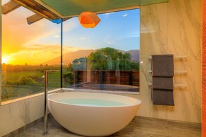A modern bathroom with a large freestanding bathtub, a glass-enclosed shower area, and a view of a sunset over a mountainous landscape.