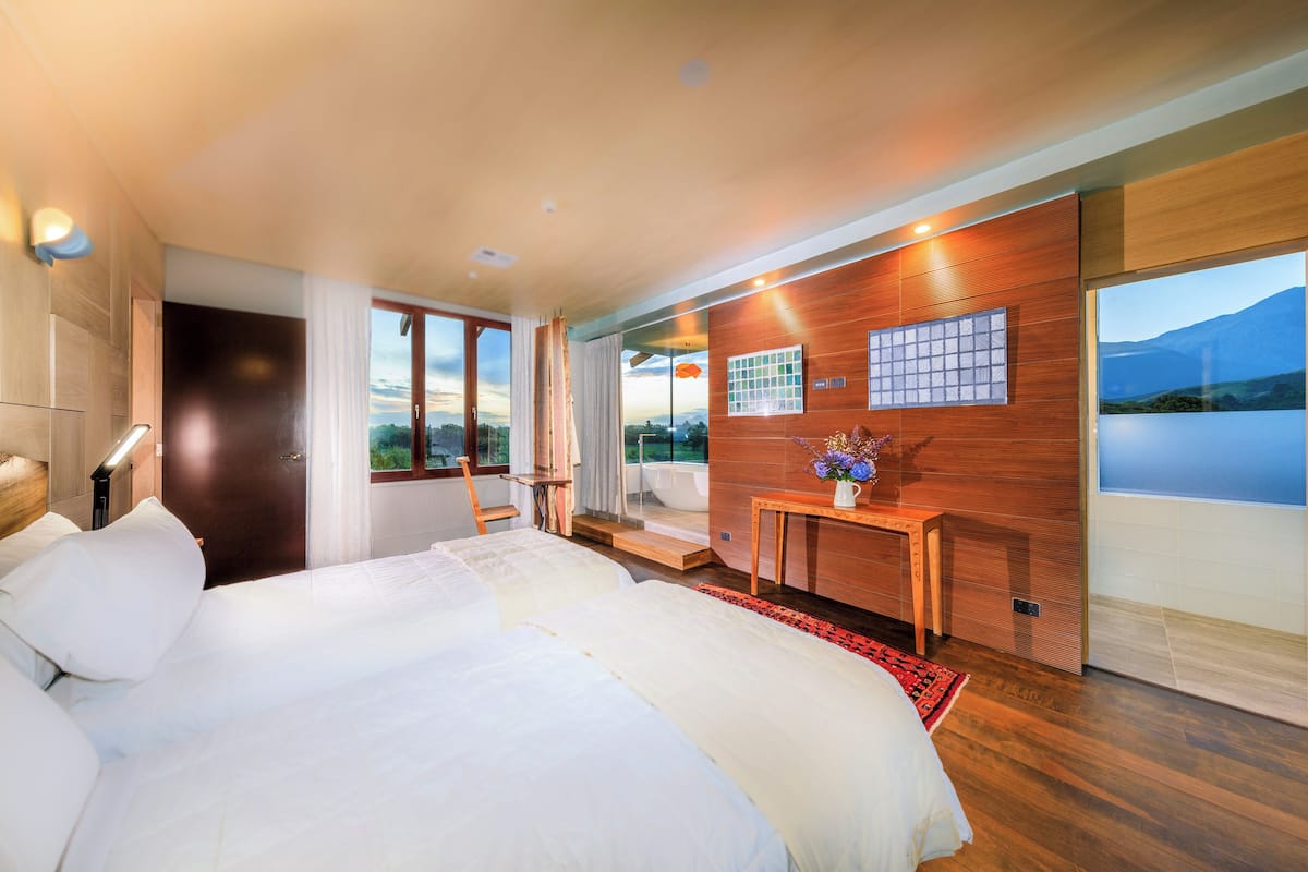 Ein modernes Hotelzimmer mit einem großen Bett, einer Wand mit Holzvertäfelung, Blick ins Freie und einem kleinen Tisch mit einer Vase.