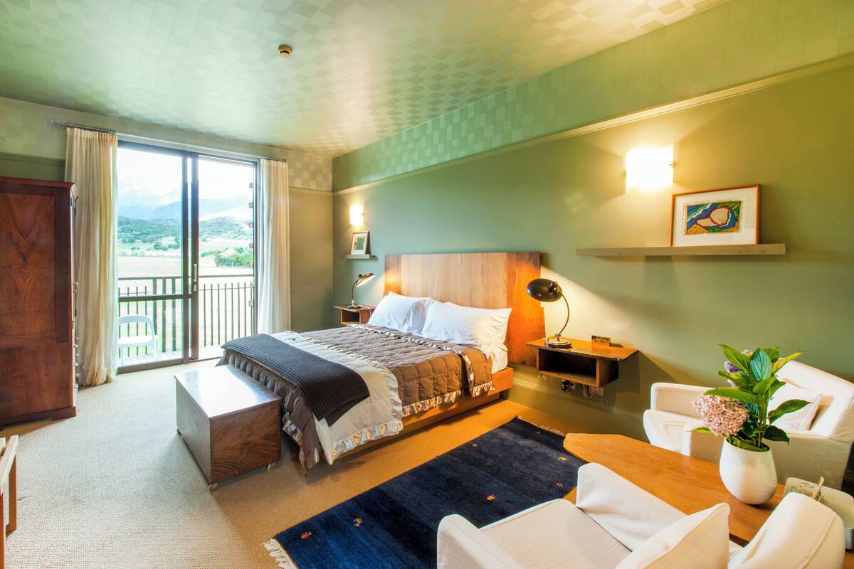 Ein Hotelzimmer mit Bett, Schreibtisch, Stuhl, Sofa, Nachttisch, Lampe, Bilderrahmen und Pflanze.