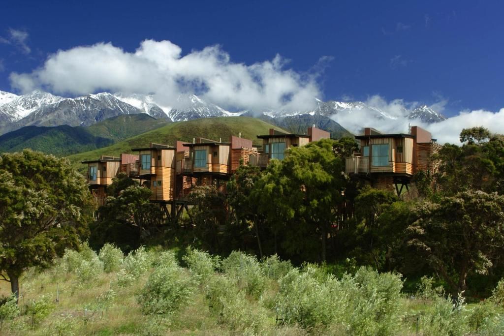 Una hilera de cabañas de madera con balcones, con montañas nevadas y un cielo azul despejado al fondo.