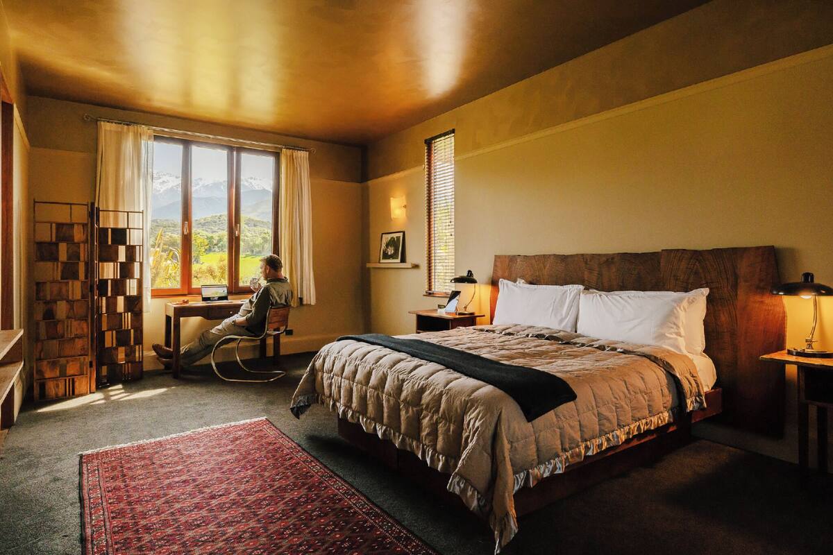 Ein Schlafzimmer mit einem großen Bett, einem Schreibtisch, einem Stuhl, einem Bücherregal und einem Fenster mit Blick auf Berge.