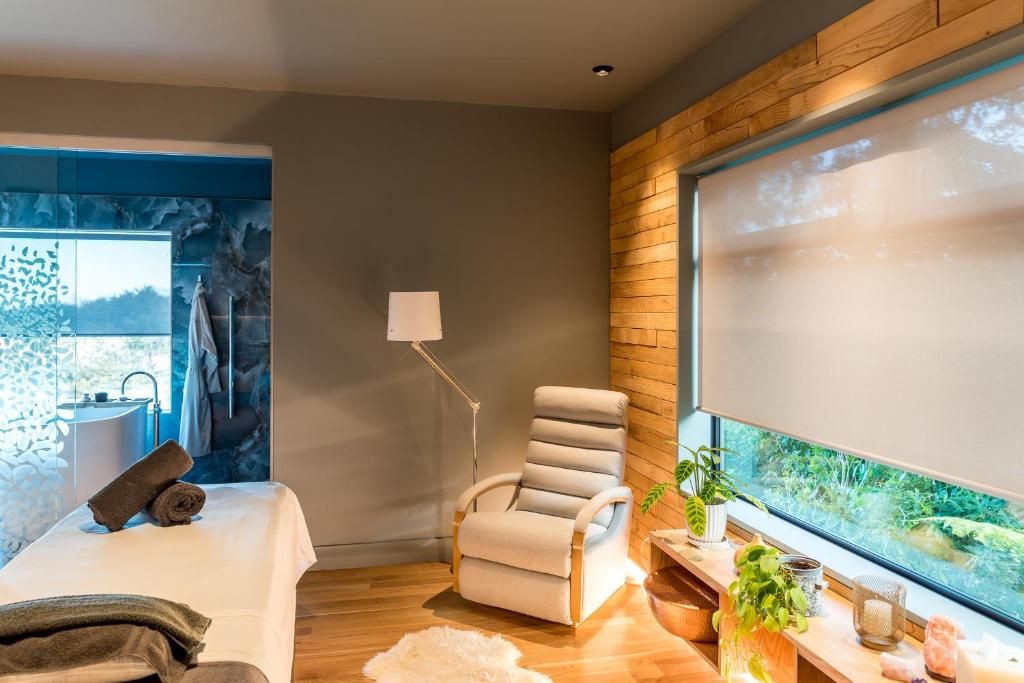 Ein moderner Wellnessraum mit Massagetisch, Liege, großem Fenster mit Blick ins Grüne und einer kleinen Innenwasserkonstruktion.