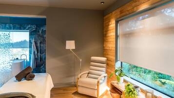Ein moderner Wellnessraum mit Massagetisch, Liege, großem Fenster mit Blick ins Grüne und einer kleinen Innenwasserkonstruktion.
