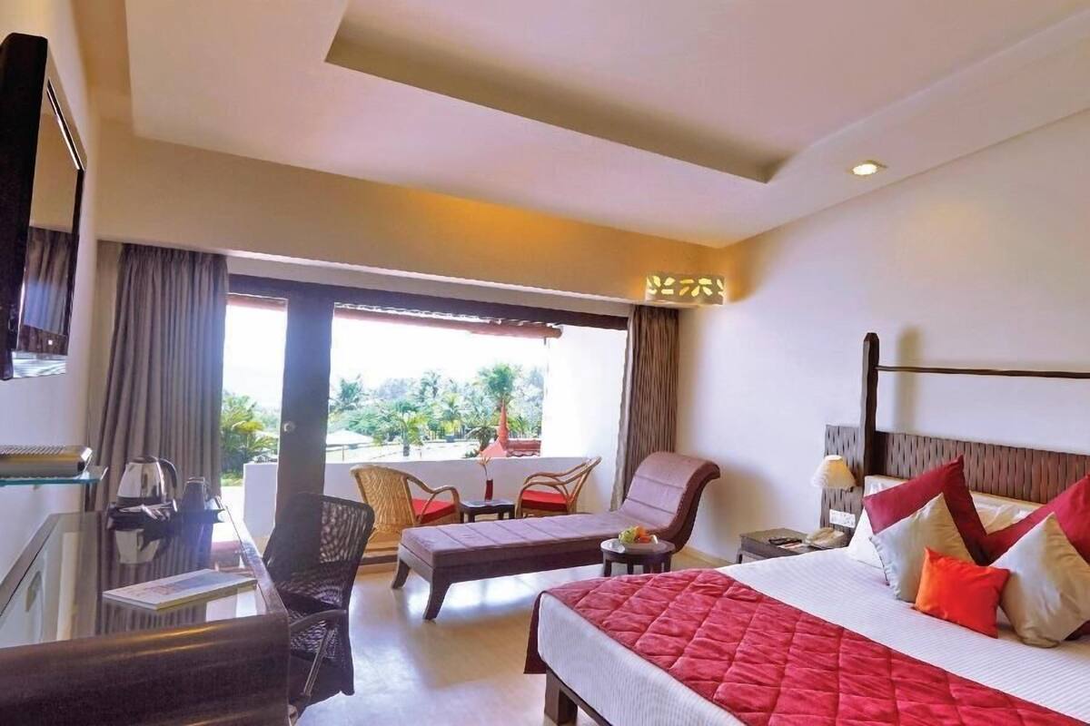 A hotel room with a bed, a desk, a chair, a small table, and a balcony with a view of greenery.
