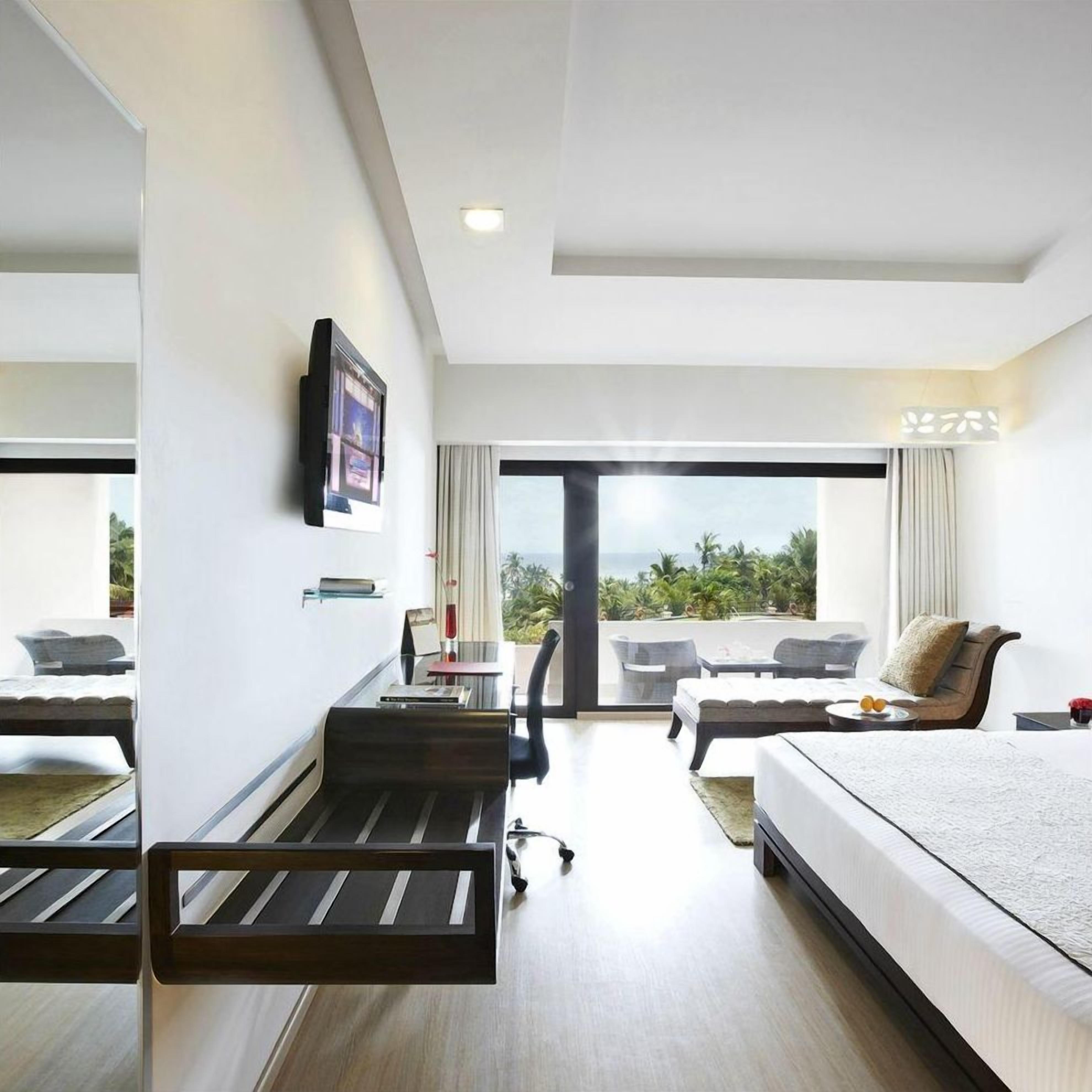 A modern hotel room with a bed, a desk, a chair, a sofa, and a large window offering a view of the outdoors.