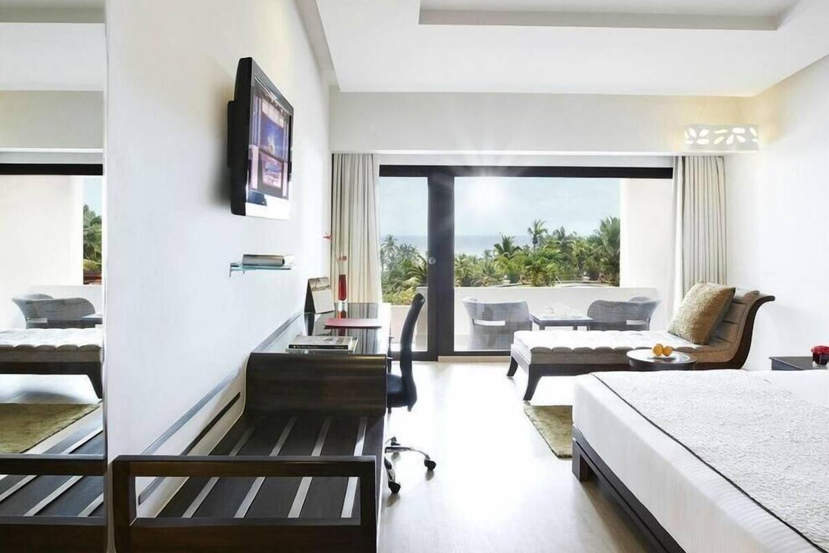 A modern hotel room with a bed, a desk, a chair, a sofa, and a large window offering a view of the outdoors.