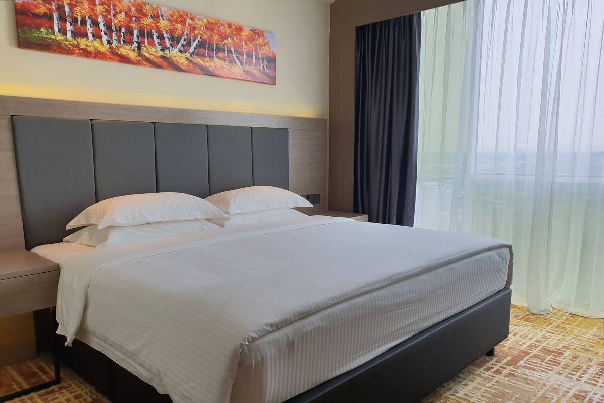 A hotel room with a large bed, a headboard, a nightstand, and a window with curtains.