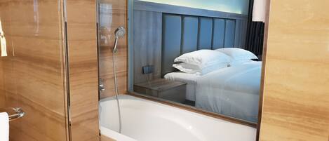 A modern bathroom with a glass-enclosed shower, a white bathtub, and a large mirror reflecting a bedroom with a bed and a painting on the wall.