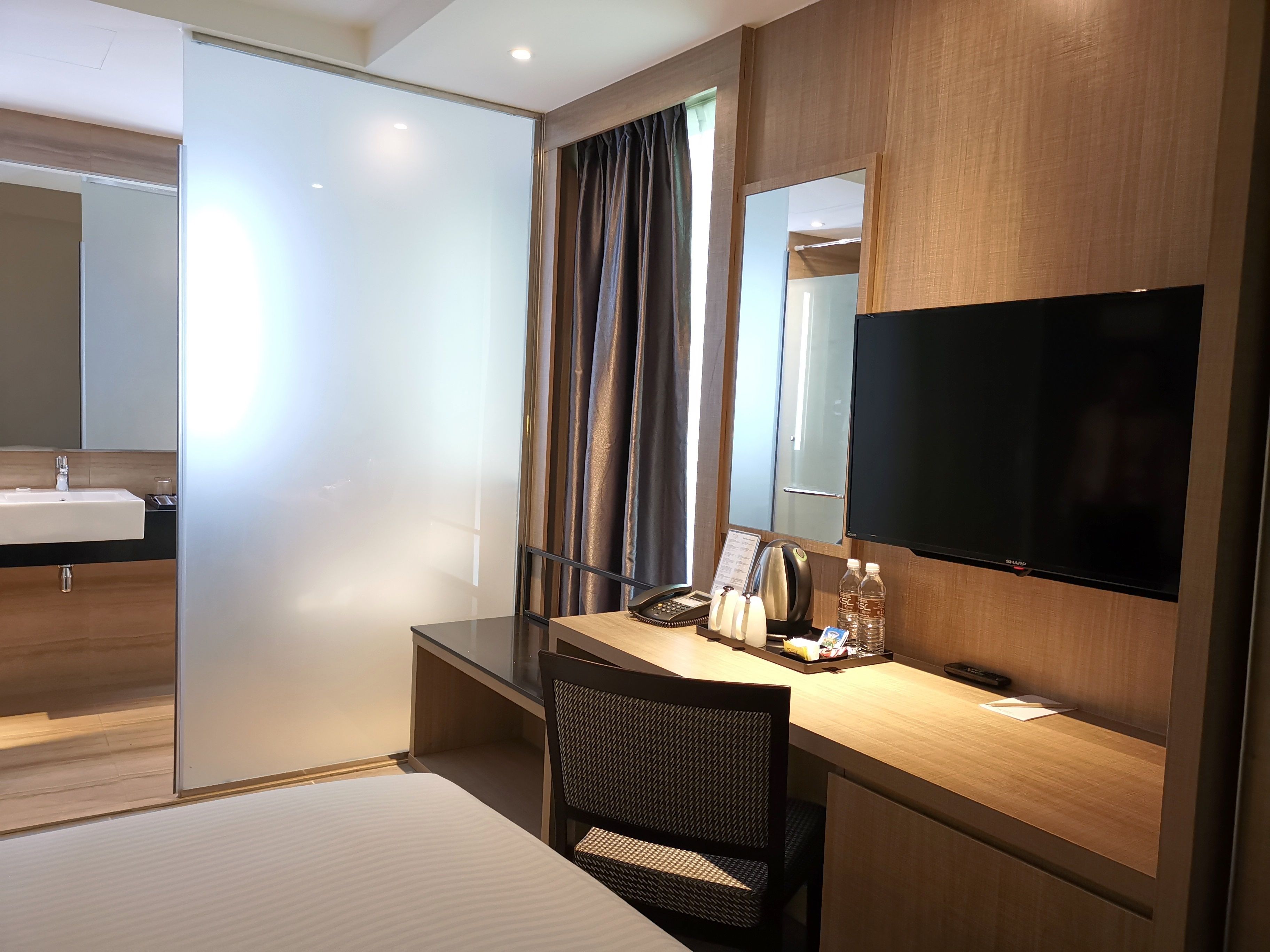 A modern hotel room with a large bed, a desk with a chair, a flat-screen TV, a mirror, a window with curtains, and a bathroom with a sink and mirror.