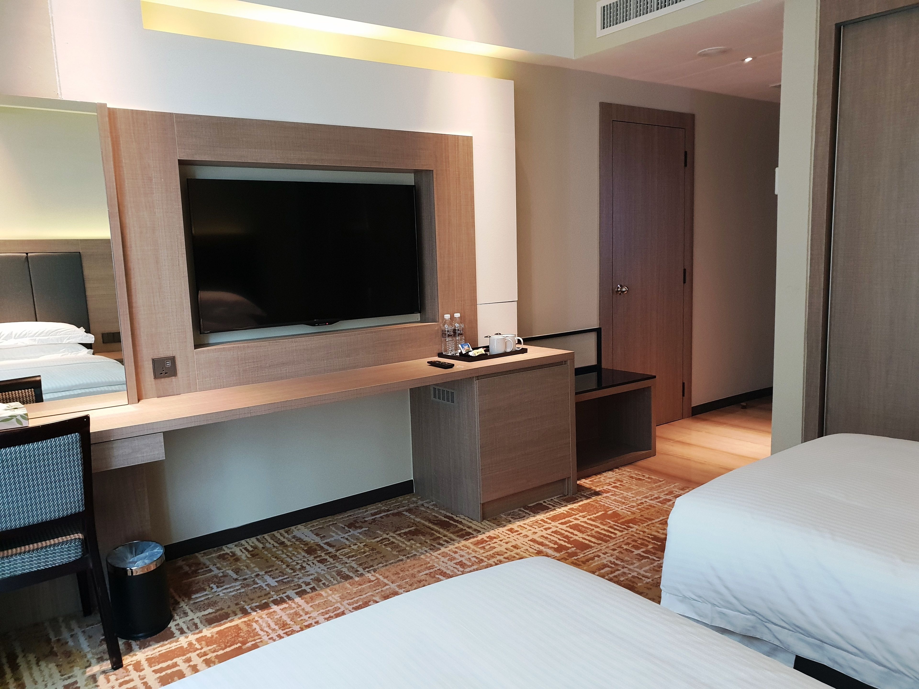 Habitación de hotel con cama, escritorio con televisor de pantalla plana, mesita con control remoto, cubo de basura y puerta.