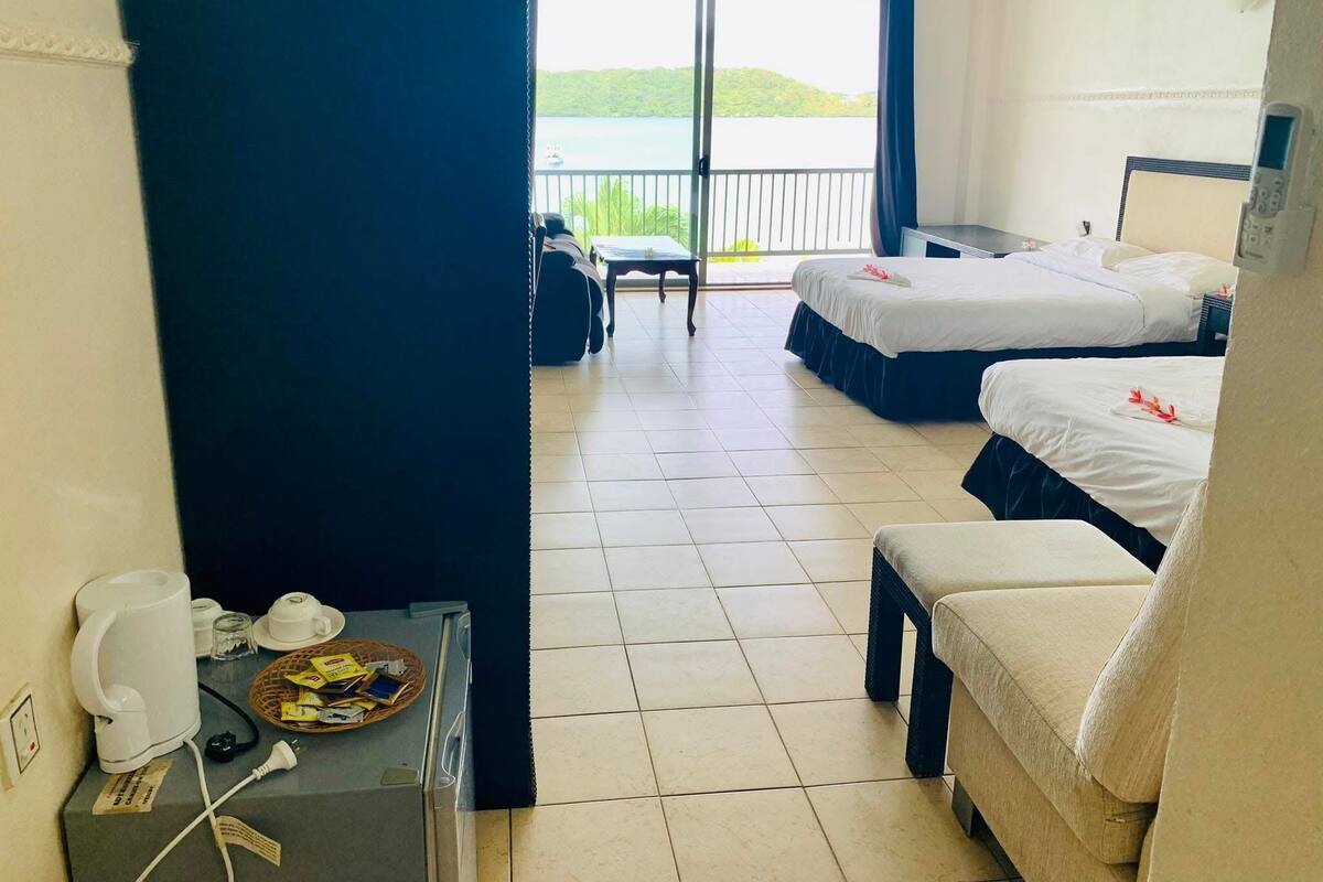 Ein Hotelzimmer mit zwei Betten, einem kleinen Tisch mit Wasserkocher und Tassen, einem Stuhl und Blick auf ein Gewässer durch ein großes Fenster.