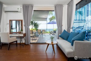A modern hotel room with a wooden desk, a sofa with blue cushions, a round table with a plant, and a balcony with outdoor furniture and a blue umbrella. - Zoneland Apartments Cani Beach Villa (Da Nang)