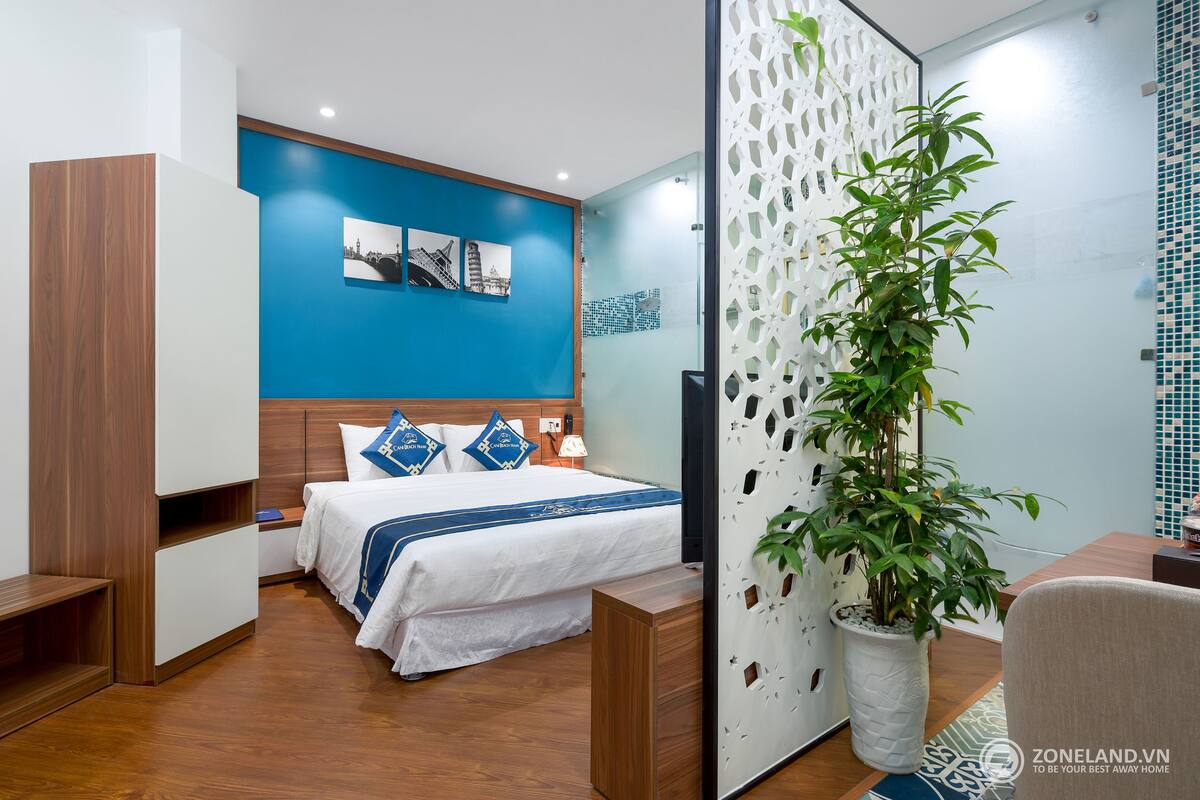 A modern hotel room with a bed, a desk, a potted plant, and a decorative wall.