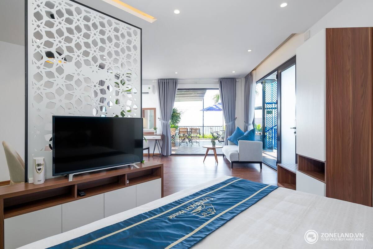 A modern hotel room with a large flat-screen TV, a bed with a blue runner, a decorative wall panel, a balcony with a view, and a wooden wardrobe.