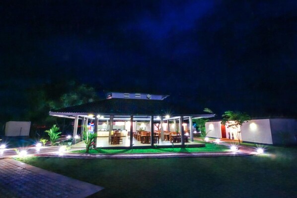 A covered outdoor dining area with tables and chairs, surrounded by a well-lit garden. - Ceyloni hotel (Inamaluwa)