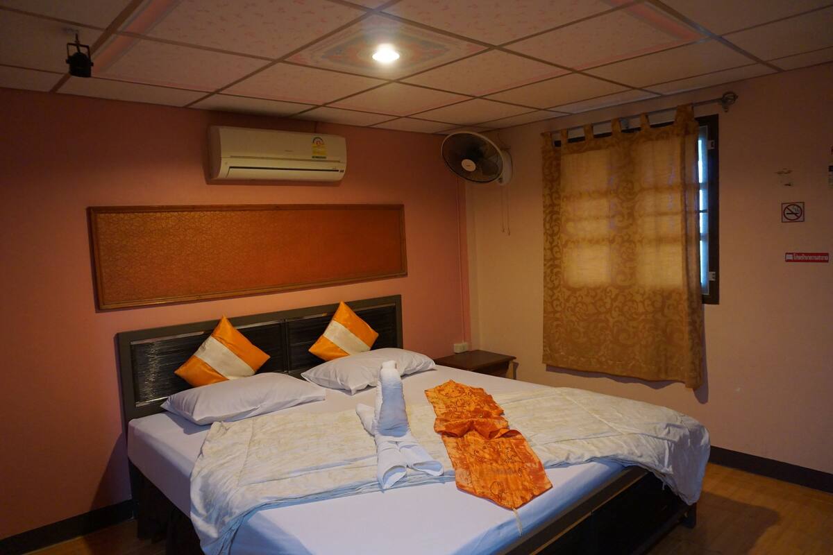 A hotel room with a bed, an air conditioner, a fan, a window with curtains, and a wall-mounted sign.