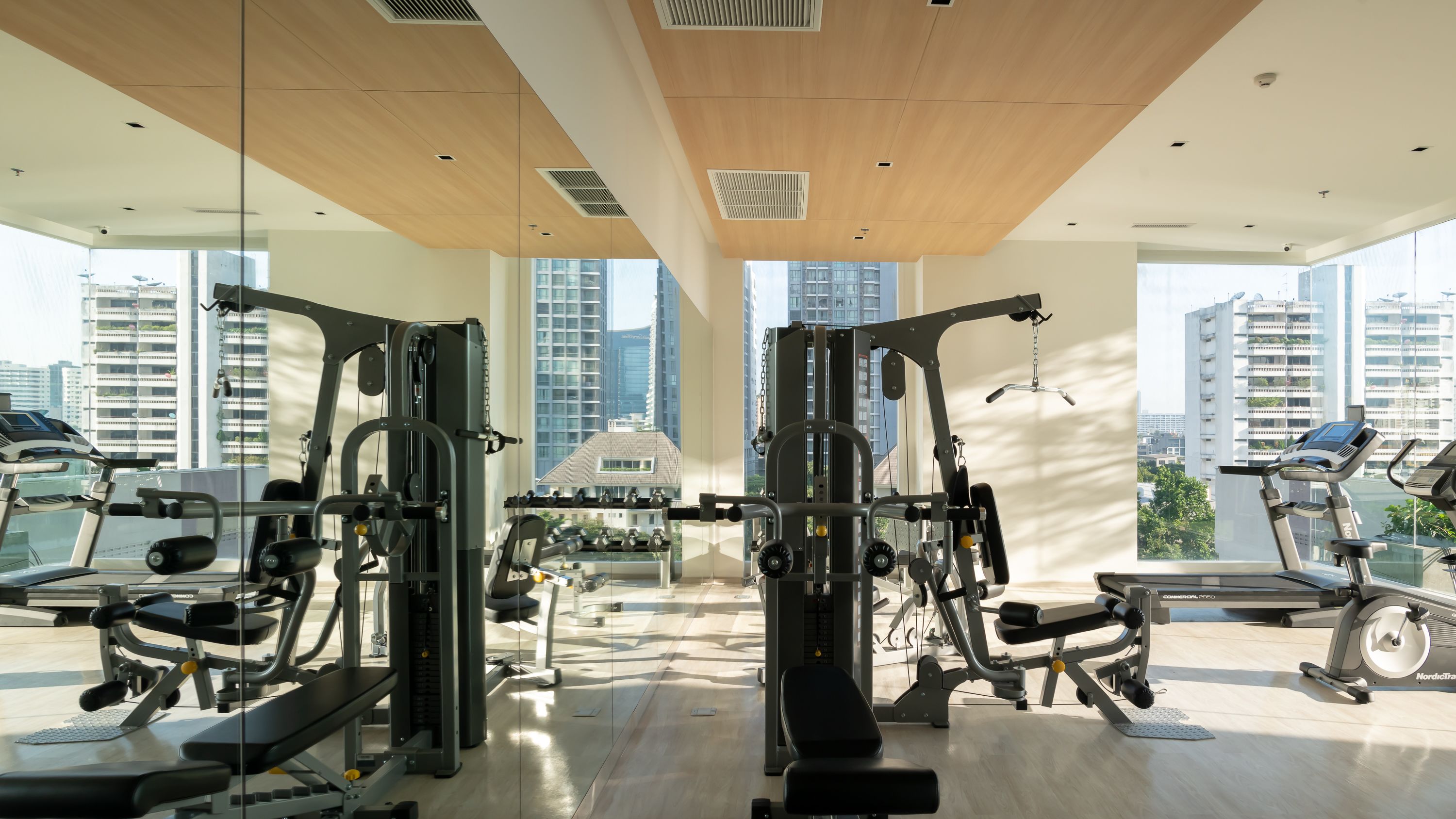 A modern gym with various exercise equipment, large windows, and a view of high-rise buildings.