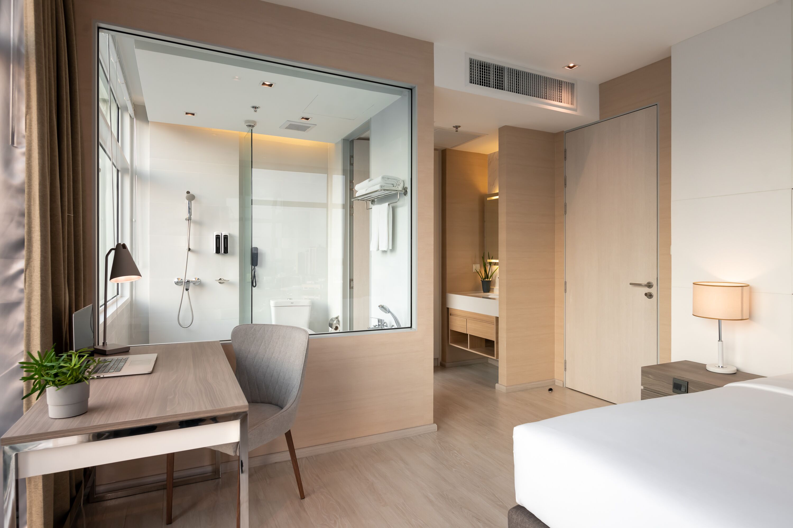 A modern hotel room with a large mirror, a desk with a laptop and a plant, a bed, a bathroom with a shower and sink, and a bedside table with a lamp.